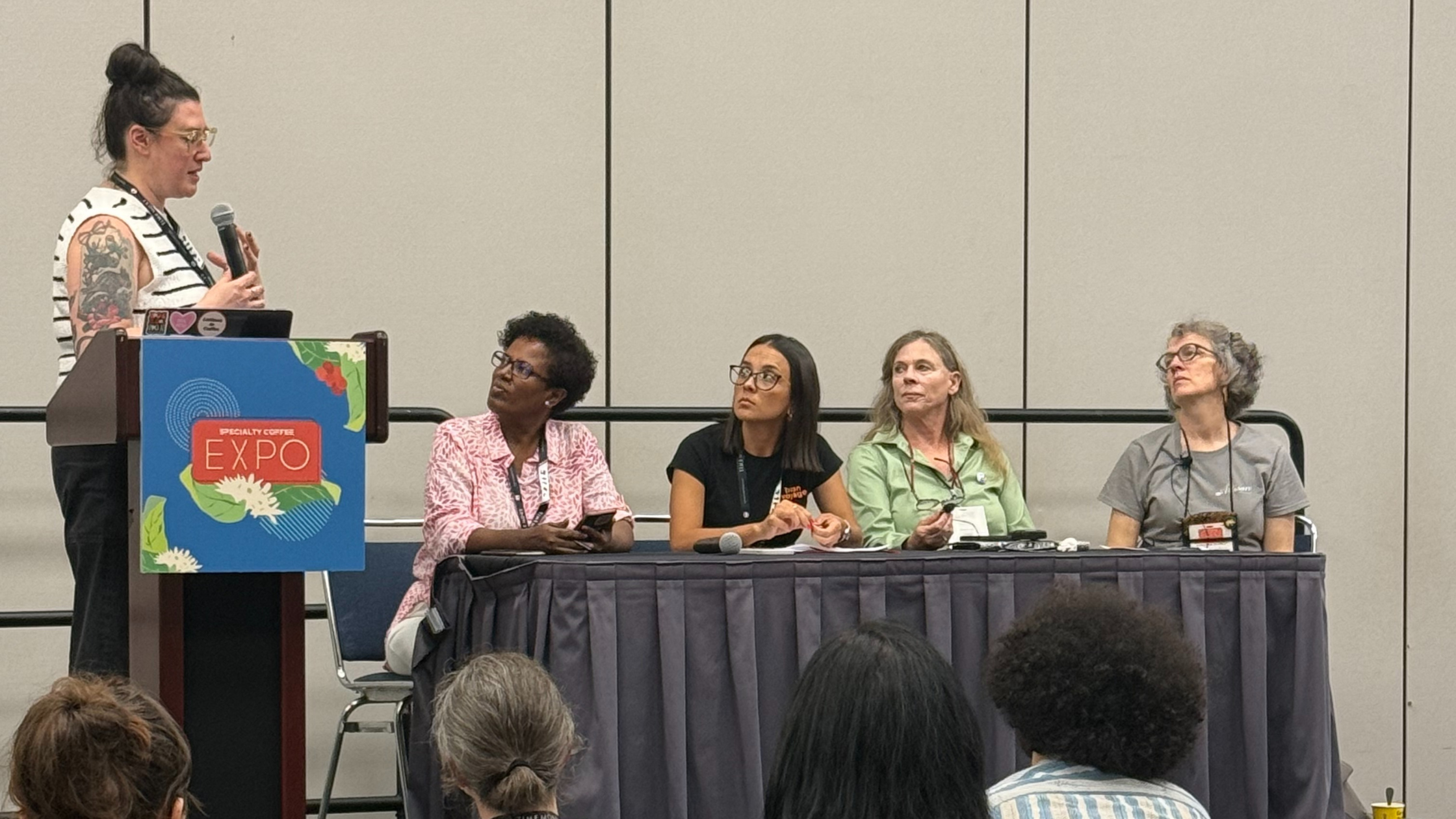 SCA Lecture Image 4 women sit at a table on a stage. One woman stands on the left behind a podium and is answering them question.